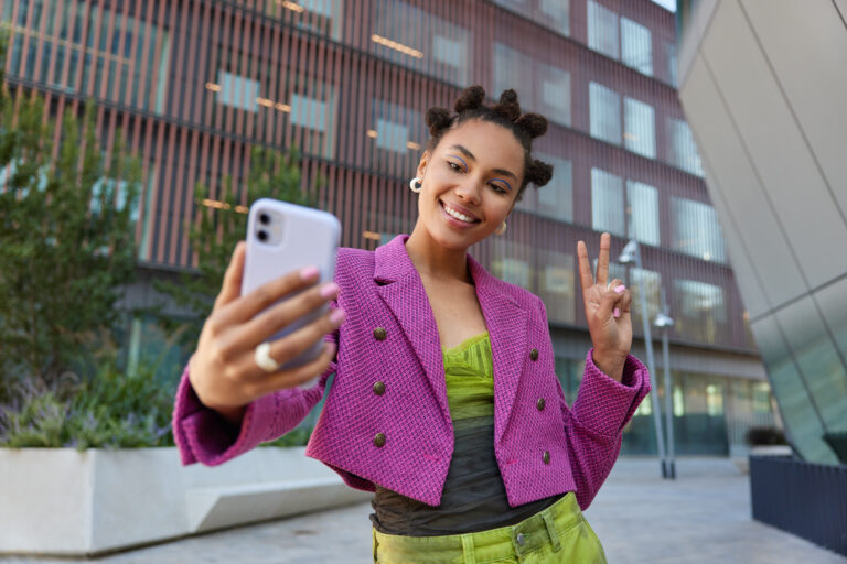 Attractive girl in fashionable outfit creats influence content shows peace sign at smartphone front camera poses for selfie smiles gladfully poses at urban place spends vacation at big city